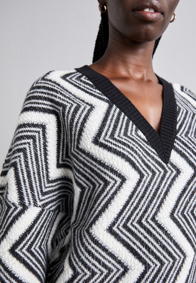 Black and white zigzag patterned sweater with a V-neck, featuring textured fabric and ribbed black neckline. Close-up view of the design.