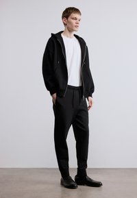 Μαύρο φούτερ με φερμουάρ και πλαϊνές τσέπες, φορεμένο πάνω από λευκό t-shirt. Συνδυάζεται με μαύρο παντελόνι φόρμας με κορδόνι και στιλβωμένα μαύρα παπούτσια.