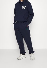 Sudadera azul marino con bolsillo frontal y logo en letras blancas, combinada con pantalones de chándal a juego que presentan un detalle de logo verde y puños elásticos.