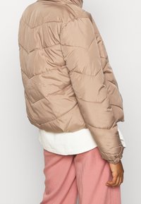 Beżowa kurtka puchowa z pikowanym wzorem, z wysokim kołnierzem i elastycznymi mankietami. Noszona na białym topie z prążkowanego materiału i różowych spodniach.