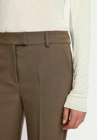 Pantaloni eleganți maro cu o strălucire subtilă, textura netedă, cu față plată și buzunare laterale, asortați cu un top lung cu mâneci lungi de un gri deschis.