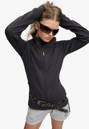 Kvinde iført sort lynlåsjakke, nittet bælte, grå shorts og store mørke solbriller med hænderne bag hovedet.