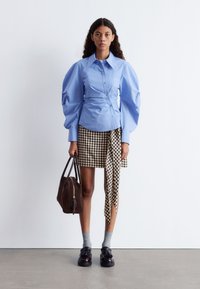 Light blue button-up shirt with puff sleeves, paired with a checkered skirt. Brown handbag and black shoes with socks complete the outfit.