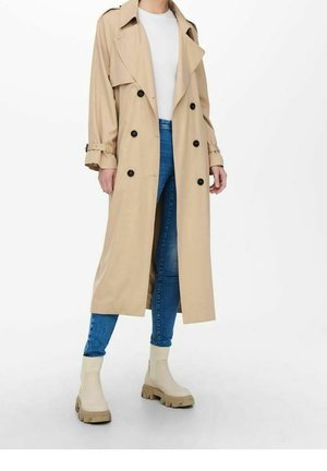 Personne portant un trench-coat beige, un jean bleu, une chemise blanche et des bottines beige, debout devant un fond blanc uni.