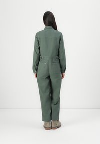 Jumpsuit en coton vert avec col, manches longues, et dos ajustable, doté de deux poches. Porté avec des bottes à cheville beiges.