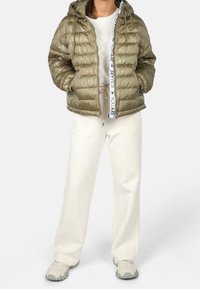 Doudoune olive brillante avec fermeture éclair à l'avant, capuche, et bande intérieure marquée ; associée à un pantalon large blanc et des baskets grises.
