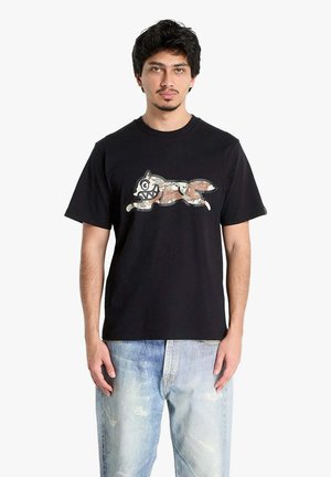 Uomo che indossa una maglietta nera con una grafica stilizzata di un animale in corsa e jeans blu sbiaditi, in piedi contro uno sfondo bianco semplice.