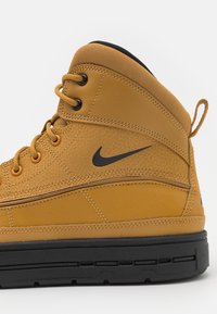 Tan läder- och mesh-högskor med svarta accenter, runda snören, texturerad yta och en logotyp nära vristen. Svart sula.