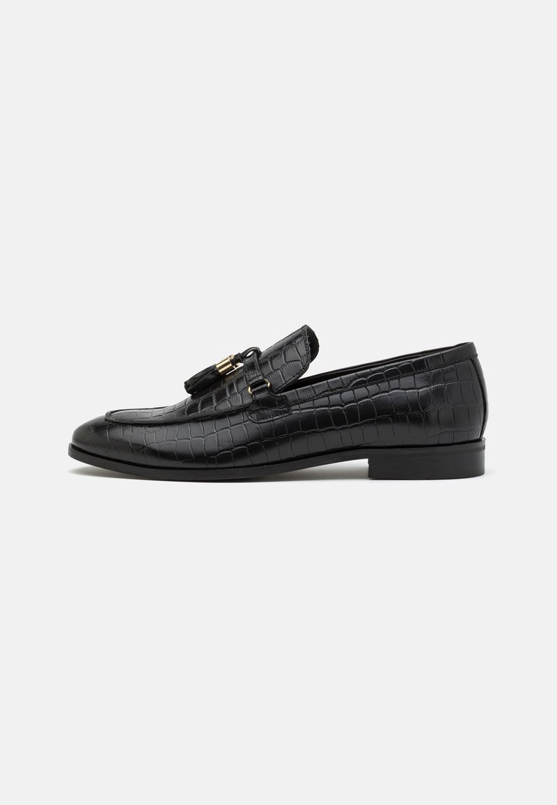 Mocasines de cuero negro con textura de cocodrilo, tacón plano, borlas decorativas y un acento de hebilla en tono dorado. Vista lateral.