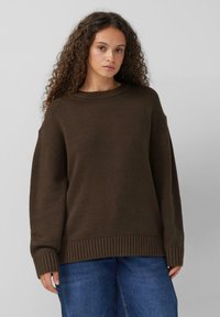 Bruine gebreide trui met een geribde zoom en manchetten, met een losse pasvorm en een brede halslijn. Draagt in combinatie met blauw denim jeans.