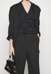 Musta räätälöity bleiseri, jonka päällä on tummasta kankaasta valmistettu, teksturoitu paita, jossa on peplum-helma. Asusteena on tilavat mustat housut ja pieni musta käsilaukku, jossa on kultaiset yksityiskohdat.