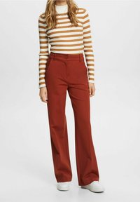 Femme portant un haut à manches longues à rayures blanches et marron, un pantalon large taille haute couleur rouille, et des baskets blanches, debout sur un fond uni.