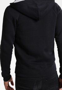 Felpa nera con una texture morbida, fodera grigia per il cappuccio, polsini elasticizzati e tasca a marsupio. Design semplice e aderente sul retro.