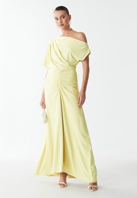 Robe jaune, décolletée sur les épaules, avec un corsage drapé, des détails froncés à la taille et une jupe évasée. Associée à des accessoires blancs.