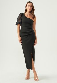 Black one-shoulder dress with a textured bodice and sheer puff sleeve. Features a side slit and fitted silhouette. Transparent heels visible.