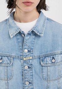 Lys blå denimjakke med klassisk krage, knappestolpe foran, to brystlommer, og en logo etikett med "REPLAY" på fronten.