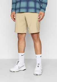 Pantalones cortos beige de corte recto, combinados con zapatillas Nike blancas y calcetines con logo. Camiseta de manga larga en color azul verdoso con un patrón de rejilla.