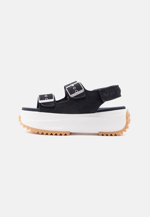 Μαύρο σανδάλι πλατφόρμα με δύο ρυθμιζόμενους ιμάντες με πόρπες, πίσω ιμάντα, χοντρή λευκή σόλα και καφέ σκισμένη πέλμα στο κάτω μέρος.