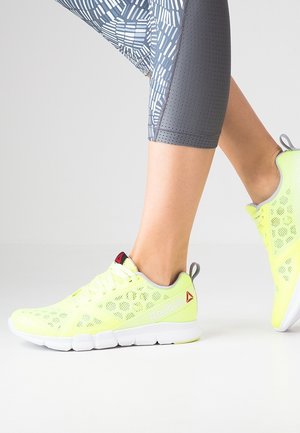 Jambes en leggings gris à motifs portant des baskets Reebok jaune vif, un pied à plat, l’autre légèrement levé.