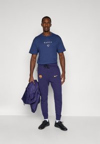 Marineblauw grafisch T-shirt met "BARCA"-tekst, paarse joggingsbroek met Nike-logo en Barcelona-badge, zwarte schoenen, een paarse jas vasthoudend.