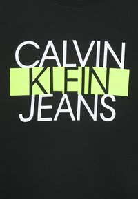 Schwarzer Sweatshirt mit fettem weißen Text "CALVIN" über "KLEIN" auf einem neon-grünen Hintergrund, mit "JEANS" in Weiß darunter. Weiche Stofftextur.