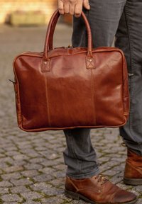 Brūna ādas portfelis ar divām rokturiem, kontrastējošiem šuvēm un ar rāvējslēdzēja aizdari. Iekļauj diskrētu zīmolu uz priekšpuses.