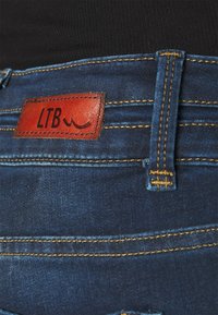 Primer plano de la cintura de unos pantalones vaqueros de mezclilla azul con una etiqueta de marca LTB de cuero marrón y costuras amarillas en el lazo del cinturón y en las costuras.