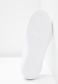 Semelle blanche de basket avec motif en zigzag, inclinée contre un mur et un sol blancs dans un cadre minimaliste.