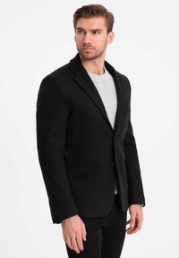 Zwarte wollen blazer met een getailleerde pasvorm, een enkele knoopsluiting, zakken en notch revers, gedragen over een lichtgrijze trui.