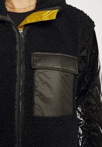 Närbild av svart texturerad fleecejacka med stor matt ficka, vadderad glänsande ärm, delvis uppdragna med gul tyg inuti kragen.