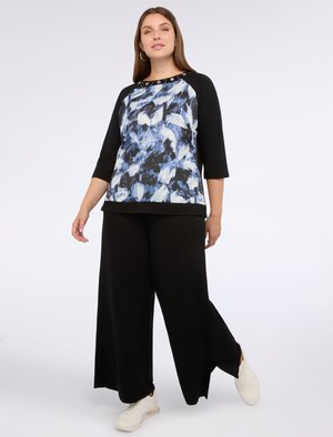 Schwarzes Top mit drei Viertelärmeln und einem blau-weißen abstrakten Muster, featuring metallic Knöpfe am Ausschnitt. Kombiniert mit weiten schwarzen Hosen.