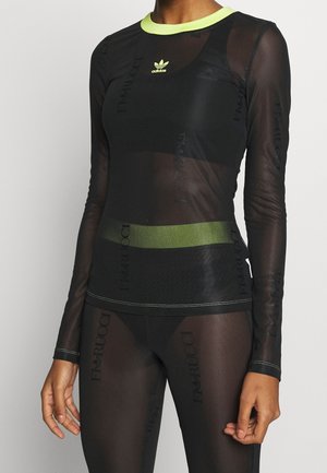 Mujer vistiendo un top de manga larga negro translúcido y leggings con detalles verdes y los logos de Adidas y Fila visibles en la tela.