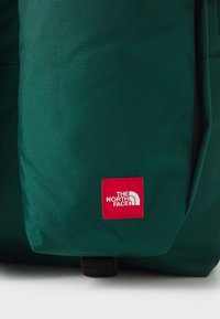 Mochila verde feita de tecido texturizado, com um emblema vermelho com texto branco e um ícone curvo, além de detalhes em metal preto.
