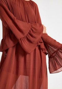 Donna che indossa un vestito lungo a maniche lunghe color ruggine con maniche a strati e svasate, realizzato in un tessuto trasparente e testurizzato.