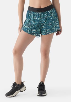 Pantalones cortos de mujer en un patrón de camuflaje azul, fabricados en un material ligero, con cintura elástica y cordón, combinados con zapatillas deportivas negras.