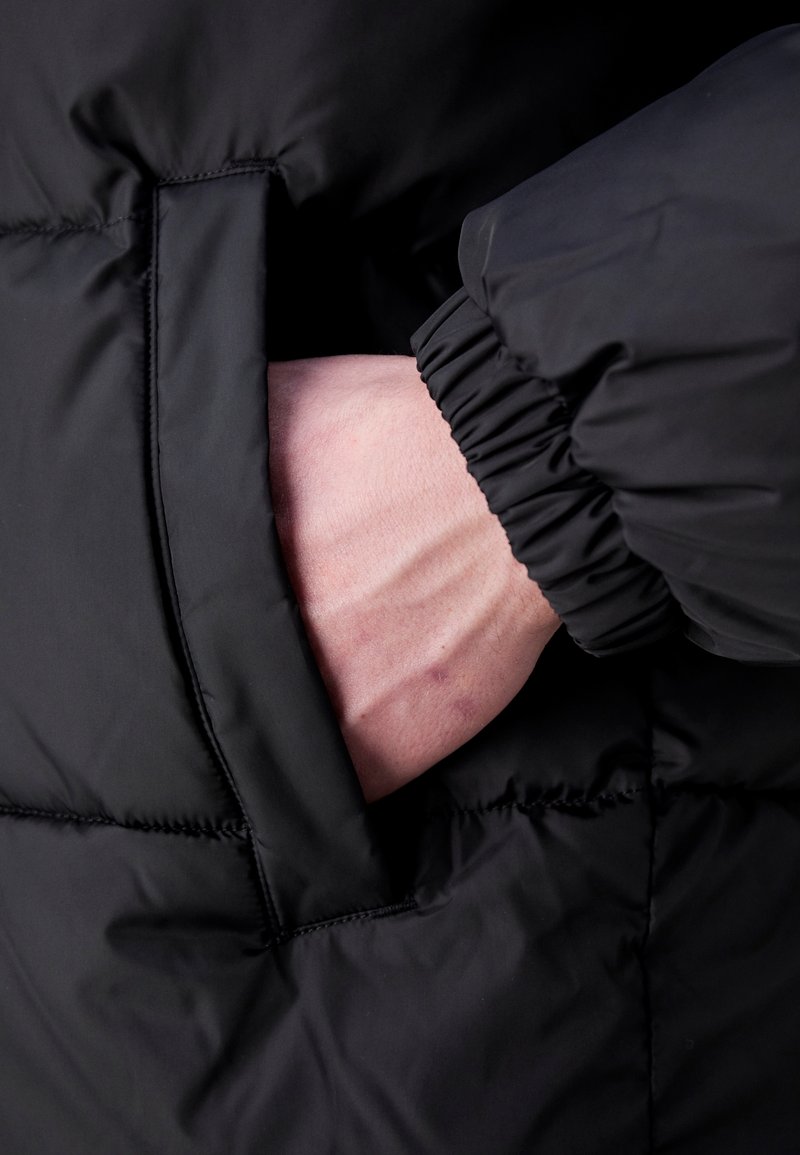 Veste matelassée noire avec un gros plan sur une main dans une poche latérale. Le tissu semble lisse et rembourré avec des poignets élastiques.