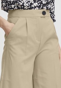 Beige High-Waist-Hosen mit Falten, Seitentaschen und einem schwarzen Knopfdetail, kombiniert mit einem floral gemusterten Oberteil. Glatte Stofftextur.