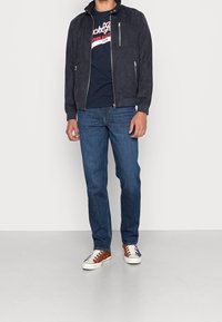 Granatowa zamszowa kurtka bomberka z kieszeniami na zamek, zestawiona z ciemnym t-shirtem z czerwonym logo, niebieskimi dżinsami oraz brązowo-białymi sneakersami.