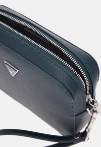 Bolso cruzado sintético en azul oscuro con forma estructurada, doble cremallera y herrajes metálicos; presenta una superficie texturizada y un acento con logo.