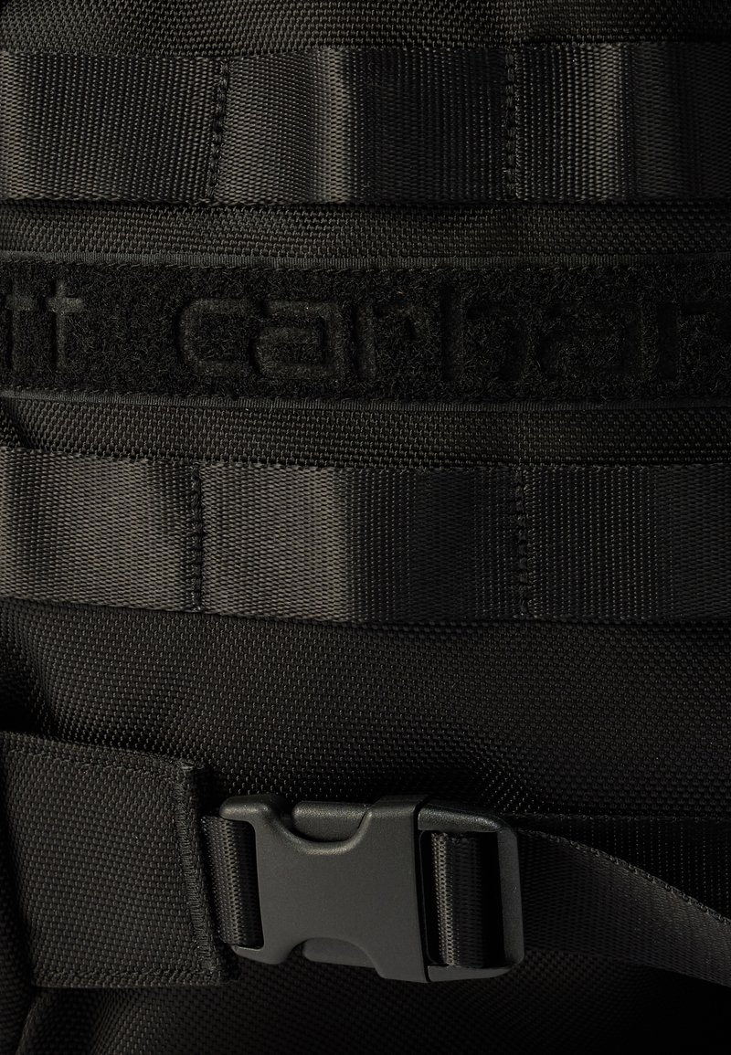 Black nylon backpack strap with textured surface, featuring a buckle and a velcro section with raised lettering for brand identification.