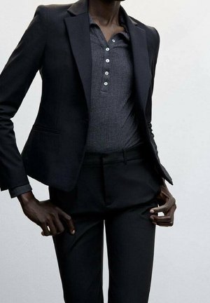 Kobieta ubrana w dopasowaną czarną marynarkę na ciemnoszarej prążkowanej koszuli zapinanej na guziki oraz czarne, dopasowane spodnie, z rękami przy biodrach.
