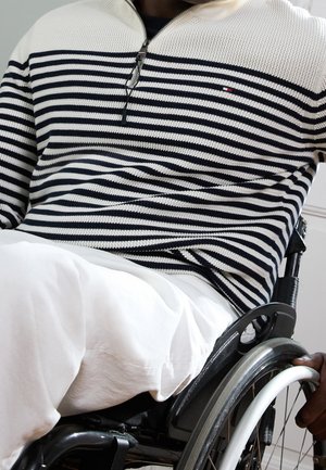 Osoba ubrana w pasiasty sweter i białe spodnie, siedząca na wózku inwalidzkim, ręka na kole wózka.