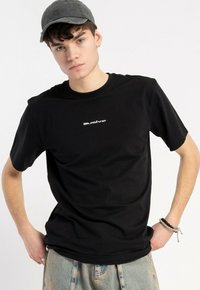 Czarny bawełniany t-shirt z białym logo "illusive" na klatce piersiowej, zestawiony z przetartymi niebieskimi dżinsami i szarym czapką.