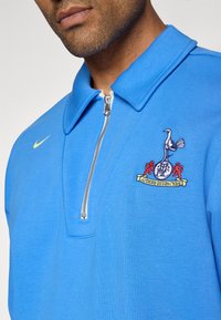 Pull-over bleu à col zippé, fabriqué en tissu lisse. Présente un logo brodé sur la poitrine et un petit swoosh Nike jaune.