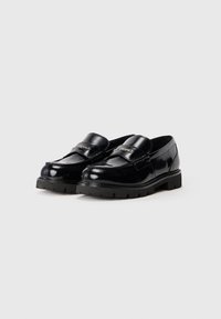 Glänzende schwarze Lederloafers mit klobigen, strukturierten Sohlen und einem markierten Metallelement am Riemen, nebeneinander auf einem weißen Hintergrund gezeigt.