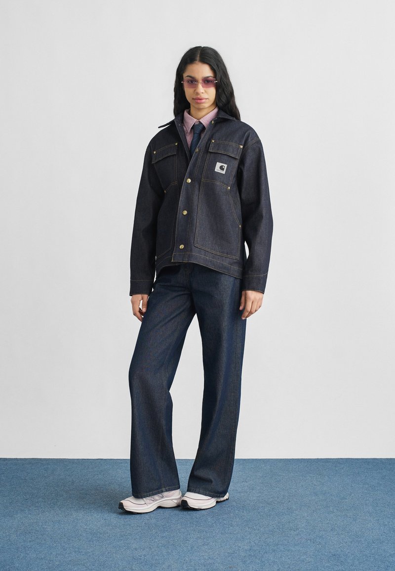 Veste en denim foncé avec poche poitrine à patch et couture dorée, associée à un jean large assorti et des baskets blanches. Comprend des lunettes de soleil teintées.