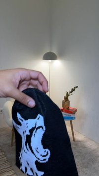 Crna kapa s bijelom slikom vuka, izrađena od pletenog materijala. Vrh ima zaobljeni oblik, s gustošću šavova i mekom teksturom.