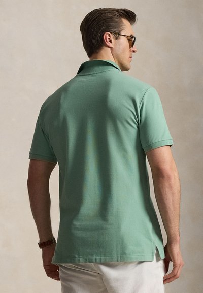 Άνδρας που φοράει πράσινο πόλο μπλουζάκι με κοντά μανίκια, άσπρο παντελόνι και γυαλιά ηλίου, εμφανιζόμενος από πίσω πάνω σε ουδέτερο φόντο.