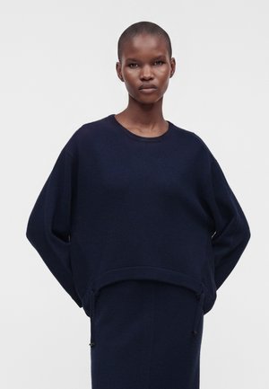 Femme aux cheveux courts portant un pull ample bleu marine foncé et une jupe assortie, debout devant un fond clair uni.