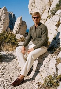 Homme portant des lunettes de soleil, un pull zippé vert, un pantalon blanc et des mocassins marron, assis sur un terrain rocheux avec de grands rochers blancs et un ciel bleu en arrière-plan.